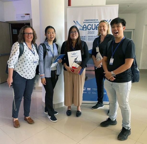 Four members of the team stand together smiling in front of an AGUA Conference banner